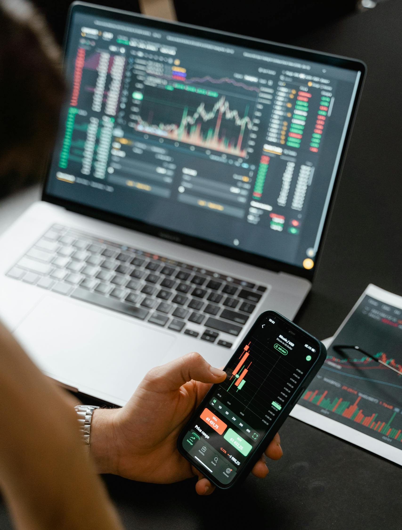 Person analyzing stock market data on a smartphone with a laptop displaying financial graphs in the background.